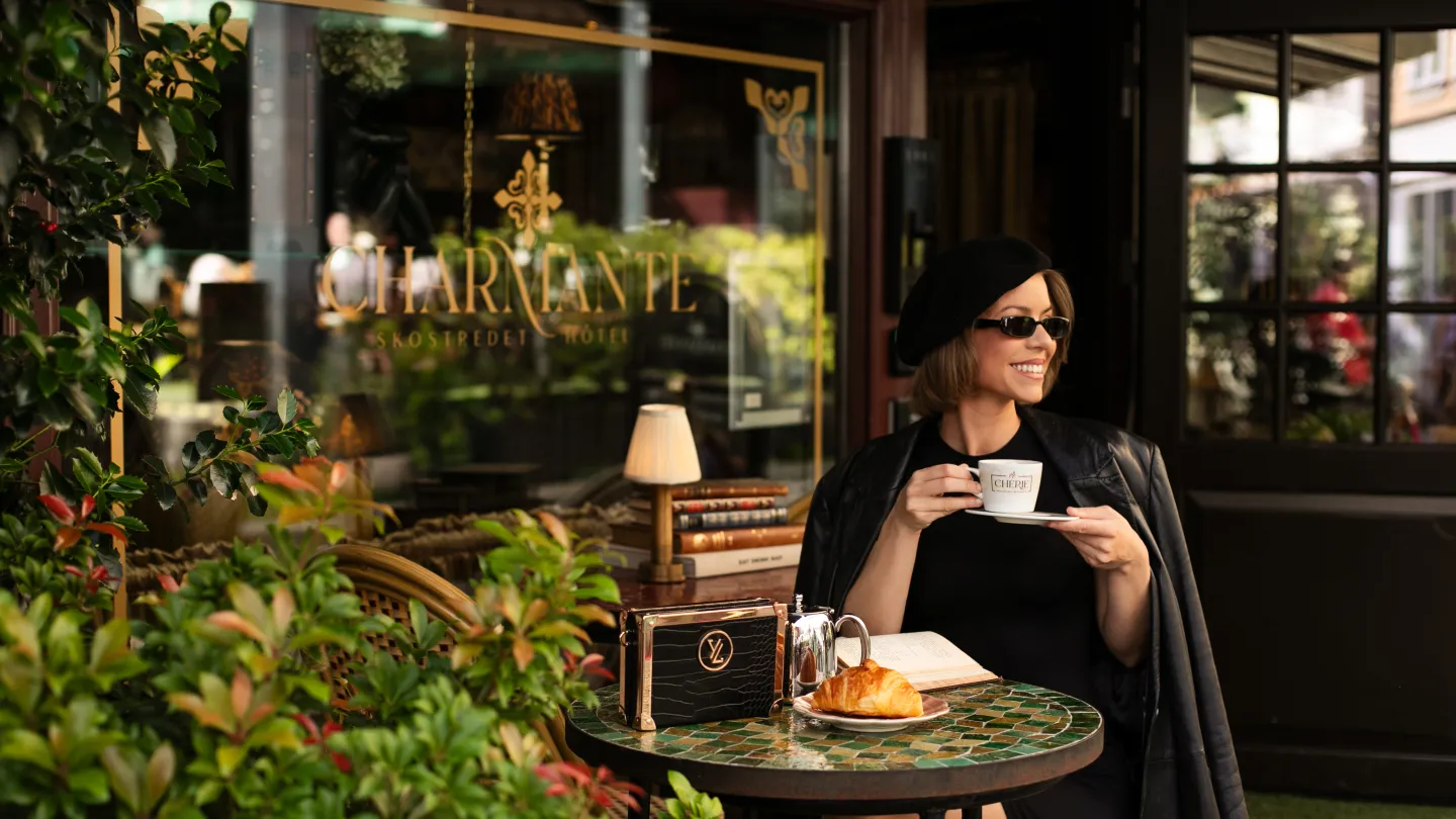 A lady enjoying her coffee outside of Charmante Skostredet Hôtel. She is smiling and holding her cup in her right hand.