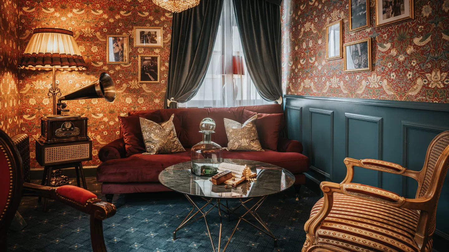 A common room in Charmante Skostredet Hôtel. A red sofa in the middle of the room with chars surrounding a cluttered table.