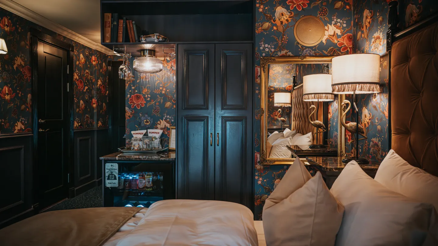 A bedroom at Charmante Skostredet Hôtel. The bed is made and in the front of the image. In the background, a black closet, a lamp with a bronze flamingo, a mirror and a mini fridge. French theme.