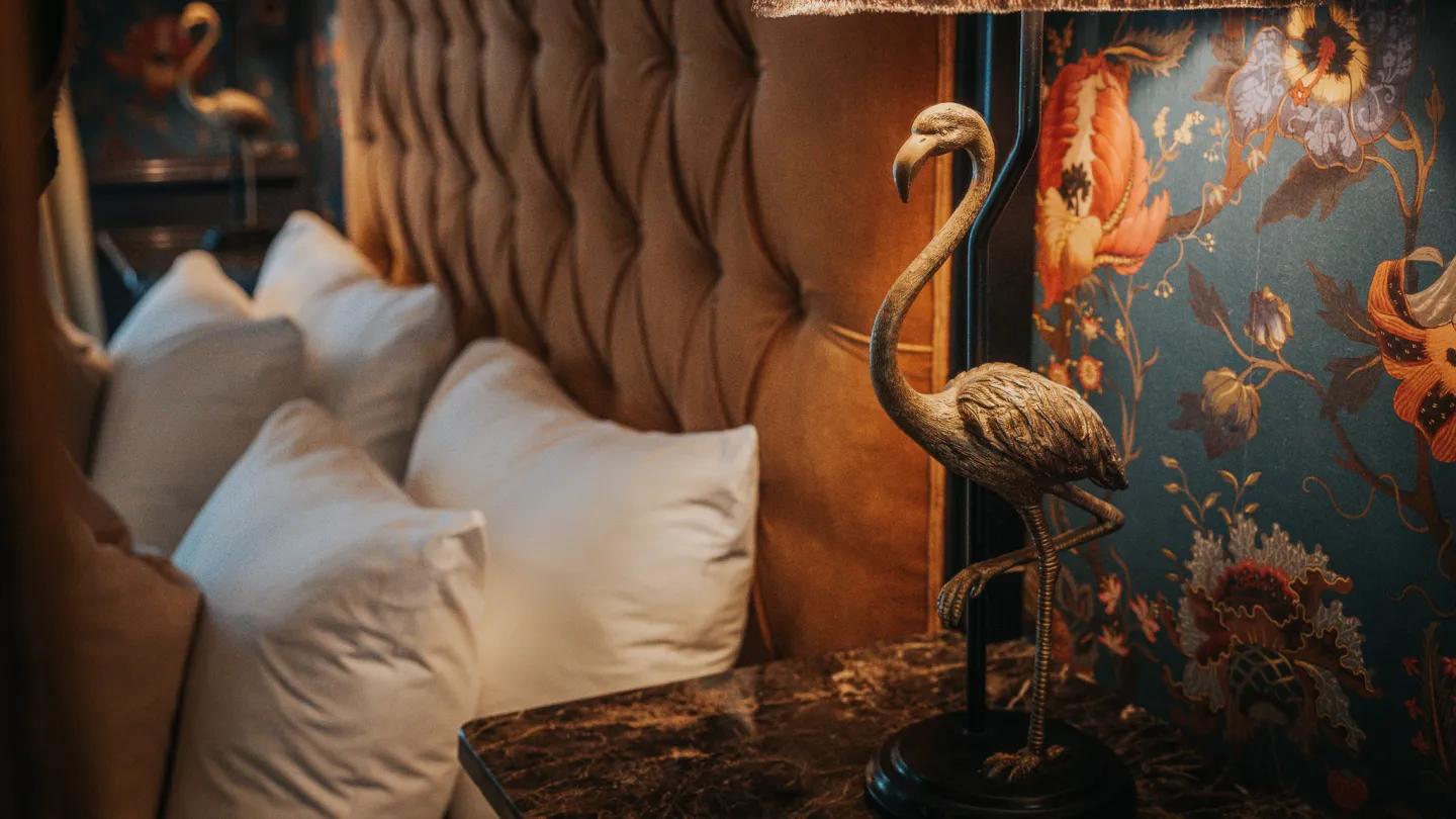 A nightstand at Charmante Skostredet Hôtel. A lamp with a bronze figure of a flamingo in front of a made bed.