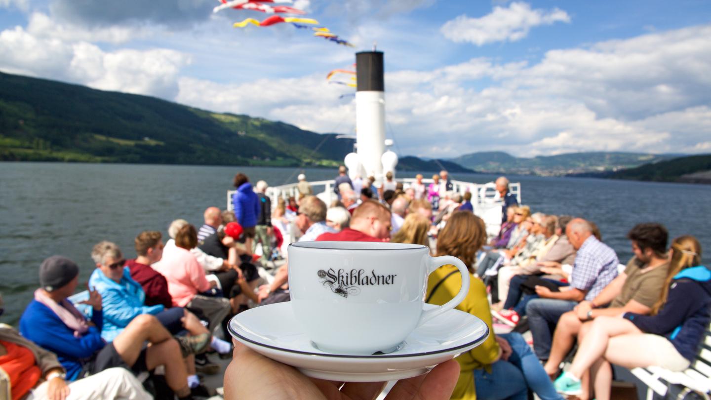 A coffee cup marked with "Skibladner" being held in front of the ships pipe and passengers